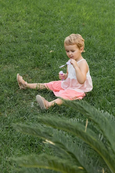 Toddler kid holding soap bubbles and sitting on grass outdoors — Stock Photo