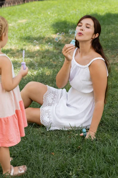 Mujer en vestido de verano soplando burbujas de jabón cerca de hija borrosa en el césped - foto de stock