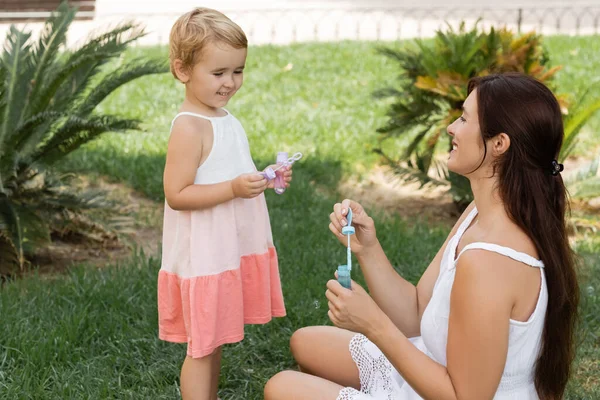 Smiling child holding soap bubbles near mom on green lawn — Stock Photo
