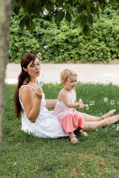 Mujer y niño pequeño soplando burbujas de jabón en el césped en el parque - foto de stock