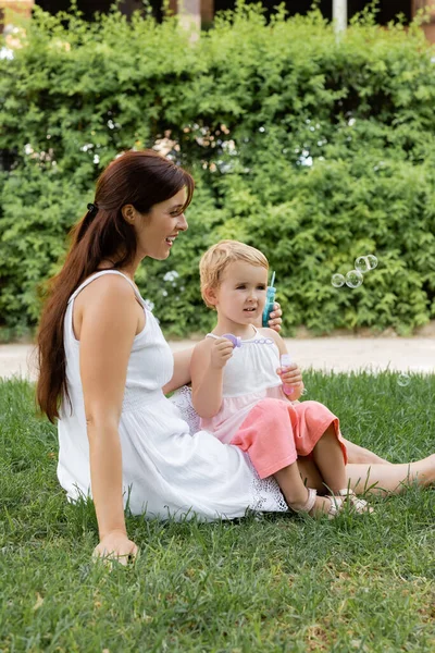 Smiling woman looking at baby daughter with soap bubbles on lawn in summer park — Stock Photo