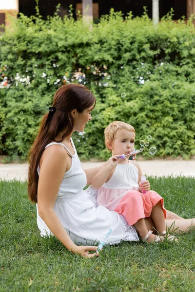 Woman looking at toddler daughter blowing soap bubbles in summer park — Stock Photo