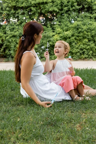 Cheerful baby girl holding soap bubbles near mom on glass in park — Stock Photo