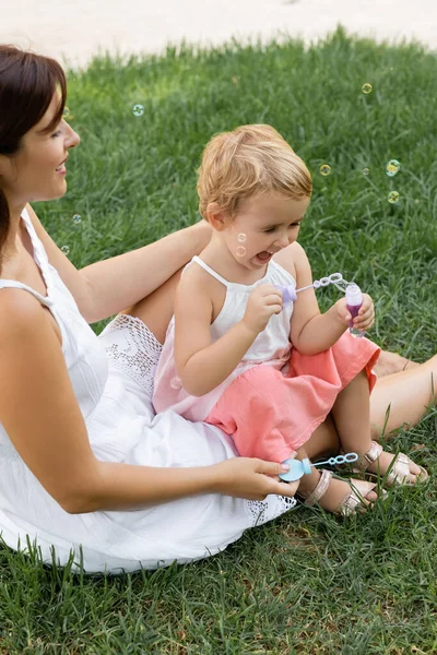 Happy mother looking at baby daughter playing with soap bubbles on lawn — Stock Photo