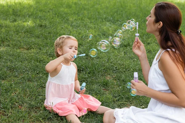 Hija en vestido soplando burbujas de jabón cerca de mamá en el césped en el parque - foto de stock