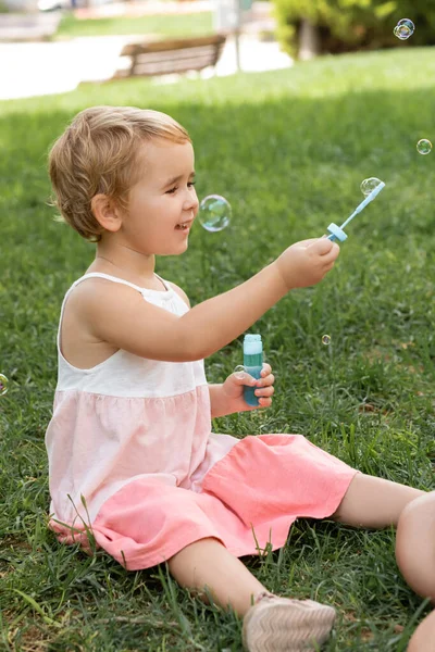 Cheerful child in summer dress playing with soap bubbles in park — Stock Photo