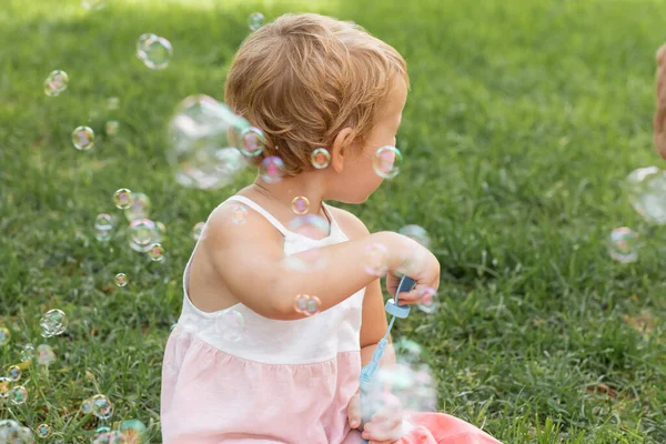 Side view of baby girl in dress holding soap bubbles in summer park — Foto stock