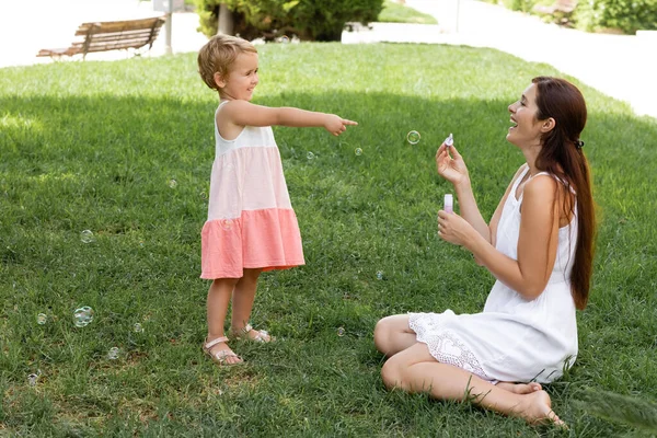 Smiling toddler girl pointing at mom with soap bubbles in park — Stock Photo