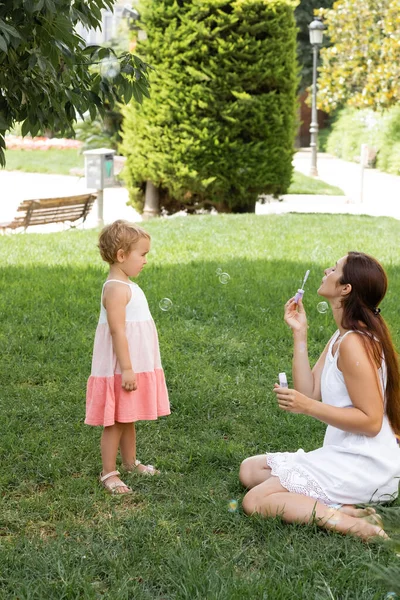 Side view of woman blowing soap bubbles near child in dress in park — Stock Photo