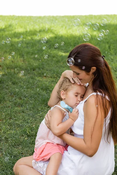 Smiling woman hugging toddler child near soap bubbles in park — Stock Photo