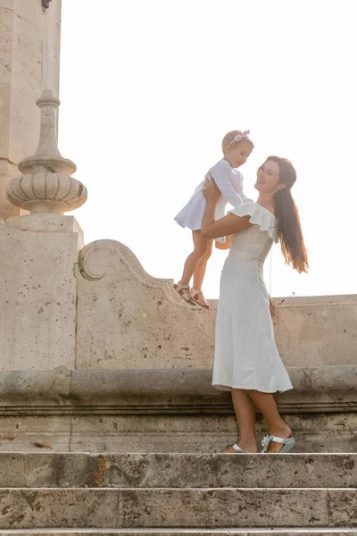 Madre morena levantando a su hija en vestido en las escaleras del puente del Mar en España - foto de stock