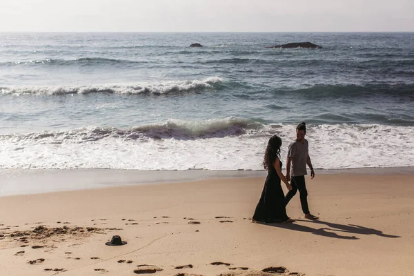 Man and woman in dress holding hands while walking on wet sand on beach near ocean — Stock Photo