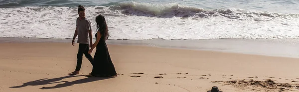 Мужчина и женщина в платье держатся за руки во время прогулки по мокрому песку на пляже возле океана, баннер — стоковое фото
