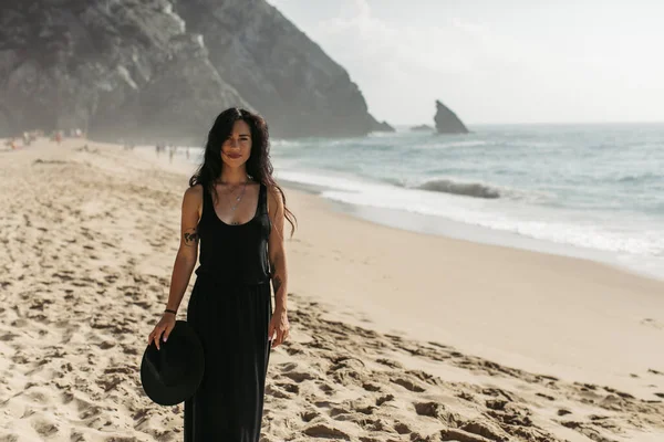 Pretty and tattooed woman in black dress holding hat while standing on sandy beach in portugal — Stock Photo