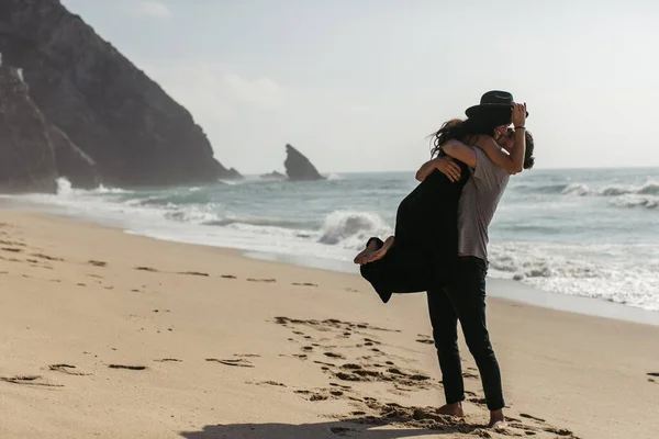 Bearded man lifting happy woman in hat and dress on beach near ocean — Stock Photo