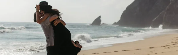 Бородатый мужчина поднимает счастливую женщину в шляпе и платье на пляже возле океана, баннер — стоковое фото