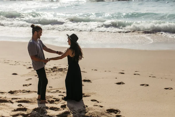 Вид збоку щасливої засмаглої пари тримає руки, стоячи на пляжі біля океану — стокове фото