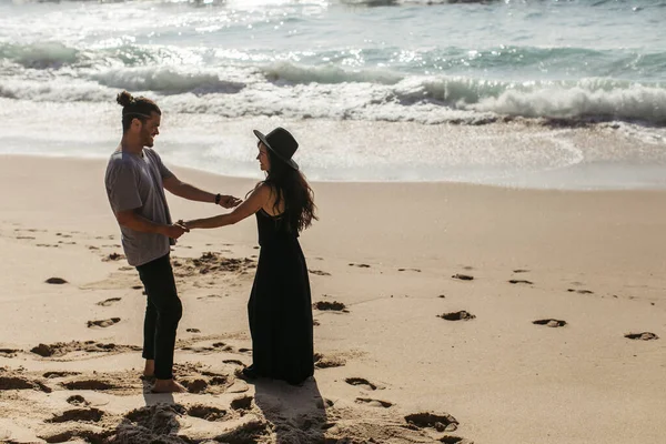 Side view of happy tanned couple holding hands while standing on beach near ocean — Stock Photo