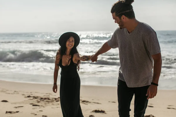 Cheerful woman in hat and black dress holding hand of boyfriend on beach in portugal — Stock Photo