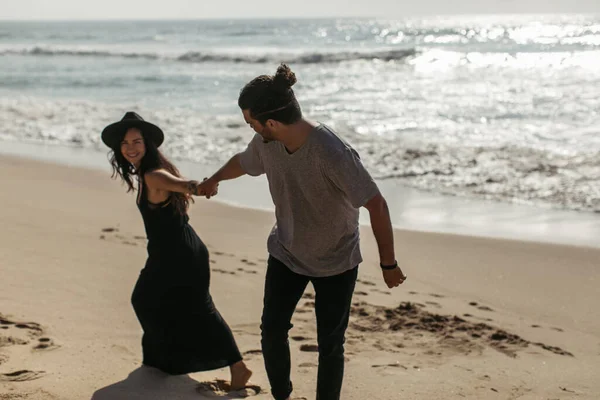 Cheerful woman in hat and black dress pulling hand of boyfriend on beach in portugal — Stock Photo