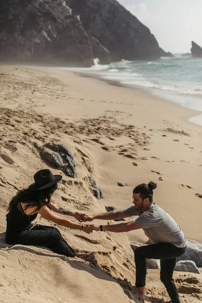 Bearded man holding hands with tattooed girlfriend in dress and hat on beach in portugal — Stock Photo