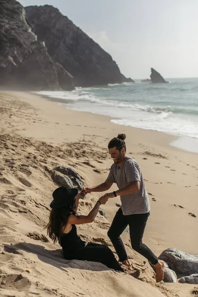 Bearded man holding hands with tattooed girlfriend in black dress and hat on beach in portugal — Stock Photo