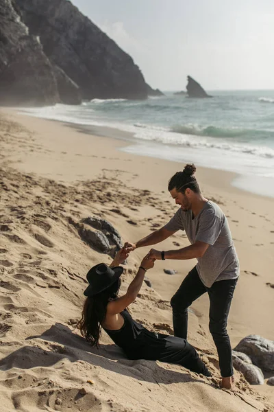 Bearded man holding hands with tattooed girlfriend in black dress and hat on sandy beach in portugal — Stock Photo