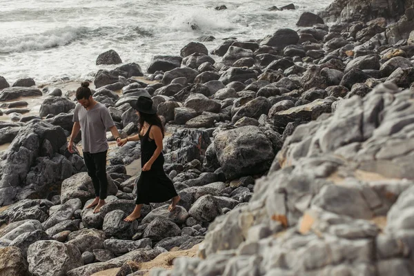 Bearded man smiling and holding hands with girlfriend in dress and hat while walking on rocks near ocean — Stock Photo