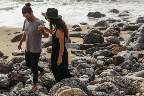 Barbudo hombre cogido de la mano con novia en vestido y sombrero mientras camina sobre rocas cerca del océano - foto de stock