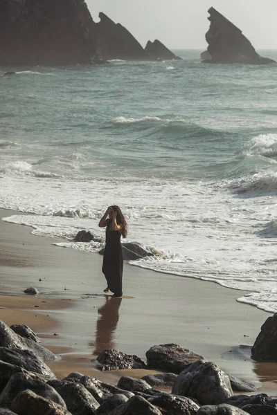 Jolie et tatouée femme en robe noire ajuster les cheveux tout en se tenant sur la plage de sable en portugais — Photo de stock