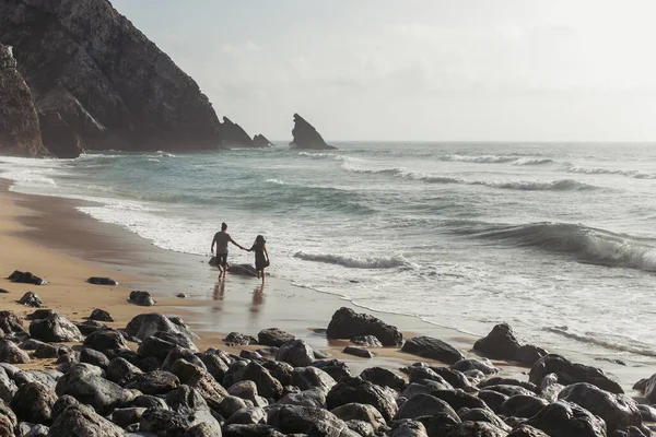 Vista trasera del hombre cogido de la mano con su novia en vestido mientras camina sobre arena mojada cerca del agua del océano - foto de stock