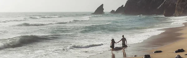 Vista trasera del hombre cogido de la mano con su novia en vestido mientras camina sobre arena mojada cerca del agua del océano, pancarta - foto de stock