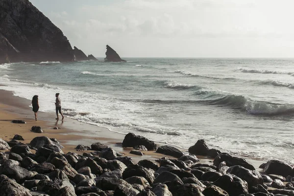 Бородатый мужчина смотрит на девушку в платье, бегущую по мокрому песку возле океанской воды — стоковое фото