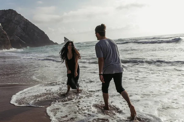 Barbuto uomo guardando felice fidanzata in abito in piedi in mare acqua — Foto stock