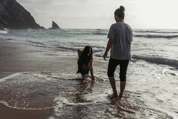 Bärtiger Mann sieht glückliche Freundin im Kleid an, die Ozeanwasser berührt — Stockfoto