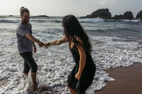 Uomo barbuto che si tiene per mano con fidanzata tatuata in abito mentre in piedi in acqua dell'oceano — Foto stock
