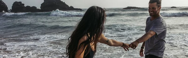 Бородатый мужчина, держась за руки с татуированной подружкой возле океана, баннер — стоковое фото