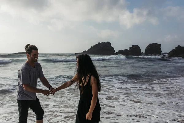 Cheerful man holding hands with tattooed girlfriend in dress while standing near ocean — Stock Photo