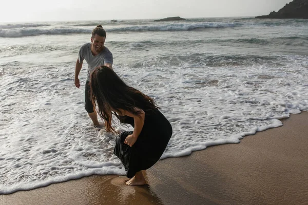 Бородатый мужчина улыбается во время веселья с подругой в платье возле океана — стоковое фото