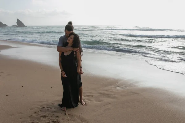 Full length of happy man hugging tattooed girlfriend in dress near ocean in portugal — Stock Photo