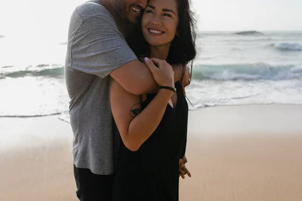 Весёлый мужчина обнимает улыбающуюся и татуированную подружку в платье рядом с океаном в Португалии — стоковое фото