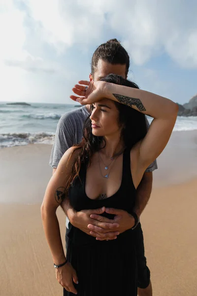 Hombre abrazando tatuado novia en vestido cerca del océano durante las vacaciones - foto de stock