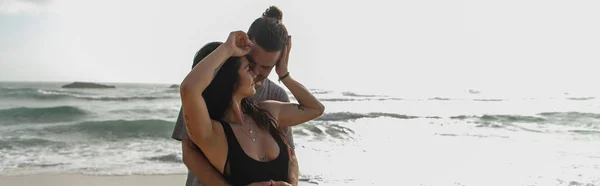 Мужчина обнимает татуированную подружку в платье и улыбается возле океана во время отпуска, баннер — стоковое фото