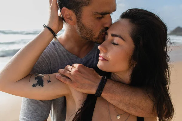Portrait of bearded man kissing tattooed girlfriend with closed eyes against sky — Stock Photo