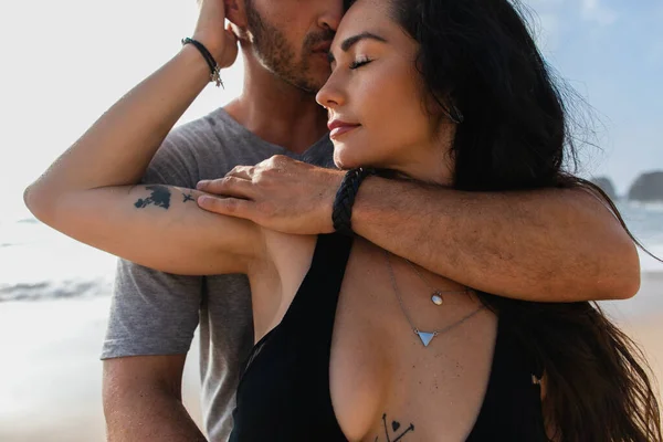 Portrait of bearded man kissing tattooed girlfriend with closed eyes near ocean — Stock Photo