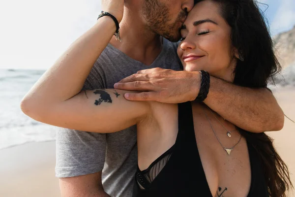 Portrait of bearded man kissing tattooed and pleased girlfriend with closed eyes near ocean — Stock Photo