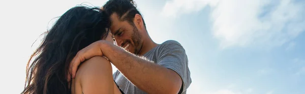 Feliz barbudo hombre abrazando morena novia contra el cielo, bandera - foto de stock