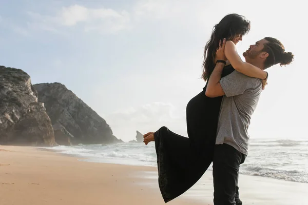 Vista lateral do homem barbudo levantando mulher em vestido na praia perto do oceano — Fotografia de Stock