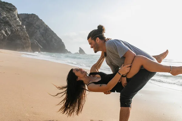 Bärtiger Mann lächelt, während er Frau im Kleid anhebt und Spaß am Strand in der Nähe des Ozeans hat — Stockfoto