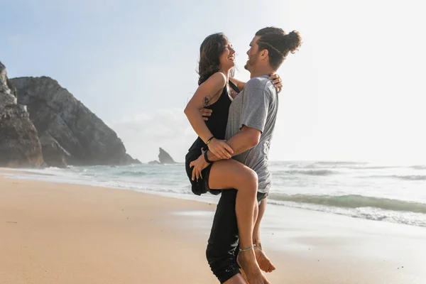 Happy bearded man lifting tattooed woman in dress on beach near ocean — Stock Photo